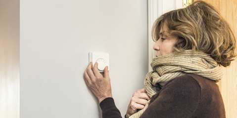A woman dressed in a warm wool jumper and scarf, adjusting a home thermostat dial.